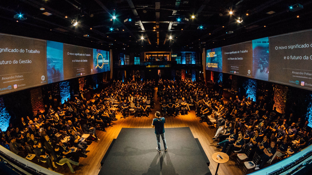 A full auditorium facing a lit stage and wide screens